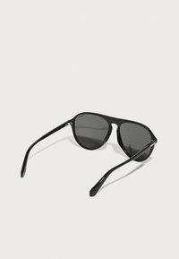 Ochelari de soare negri cu lentile întunecate, ramă rotundă și brațe negre elegante. Au un hardware minim și un finisaj lucios.