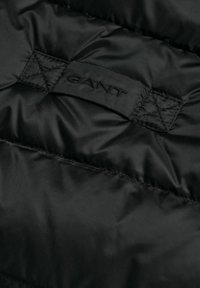 Zwarte pufferjack stof met een doorgestikt textuur. Kenmerkt zich door een zwart geborduurd "GANT" label en stikdetails. Glad en glanzend materiaal.