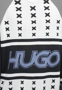 Pleteno jopico z črno-bele barvne sheme, z izrazitim logotipom "HUGO" v modri barvi in naglasi v obliki križa. Oblika z V-izrezom.