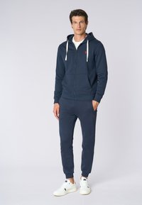 Felpa con zip di colore blu navy, dotata di cappuccio con cordino e logo rosso, abbinata a pantaloni joggers coordinati e sneakers bianche. Tessuto in morbido cotone misto.