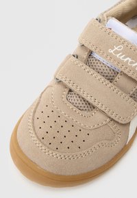 Pantof din piele întoarsă bej cu panouri din plasă, două barete cu velcro, toe box perforeat și talpă de cauciuc maro. Prezintă detalii accentuate albe.