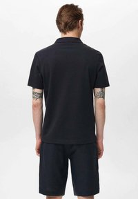 Svart herr-polo med korta ärmar och krage, tillverkad av mjukt tyg. Bärs med mörka shorts, med tatueringdetalj på armen.