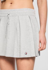 Jupe plissée grise avec taille à cordon portée par une personne, présentant un petit emblème en forme d'étoile près de l'ourlet du côté gauche.