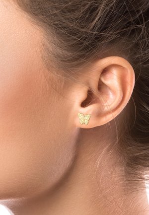 Gros plan sur l'oreille d'une femme portant une petite boucle d'oreille en forme de papillon en or, avec des cheveux bruns tirés en arrière.