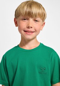 Grön bomullströja med rund hals och korta ärmar. Har en broderad logotyp på vänster bröstområde. Slät textur.