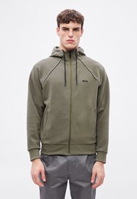 Oliwkowa bluza z kapturem na zamek, z regulowanym sznurkiem w kapturze, kontrastowymi białymi przeszyciami wzdłuż szwów oraz dwoma przednimi kieszeniami. Logo "BOSS" w kolorze czarnym.