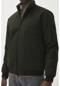 Dunkelgrüne Bomberjacke mit einem Reißverschluss vorne, enganliegenden Manschetten und einer neutralen inneren Schicht. Glattes, wasserabweisendes Material.