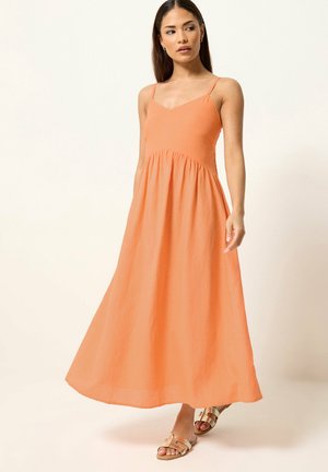 Vrouw draagt een mouwloze, enkellange oranje jurk met dunne bandjes en beige sandalen, staand tegen een effen lichte achtergrond.