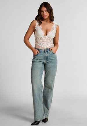 Vrouw staat met handen in haar zakken, draagt een wit kanten mouwloos topje en een lichtblauwe spijkerbroek met hoge taille tegen een effen achtergrond.