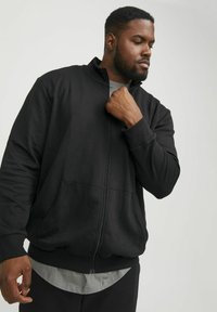 Sudadera negra con cremallera y un alto cuello, con bolsillos frontales y puños acanalados. Llevada sobre una camiseta gris, que presenta una tela suave y texturizada.