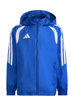 Junge in blauer Jacke und schwarzen Hosen steht mit einem Fuß auf einem Fußball, lächelt und schaut zur Seite.
