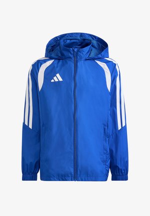 Junge in blauer Jacke und schwarzen Hosen steht mit einem Fuß auf einem Fußball, lächelt und schaut zur Seite.