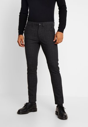 Man draagt een slim-fit donkergrijze geruite broek, een zwart overhemd met lange mouwen, zwarte sokken en zwarte veterlaarzen, staand op een witte vloer.