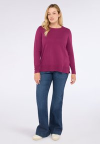 Mujer de pie vistiendo un suéter morado de manga larga, jeans de pierna ancha azules y zapatillas blancas frente a un fondo blanco liso.