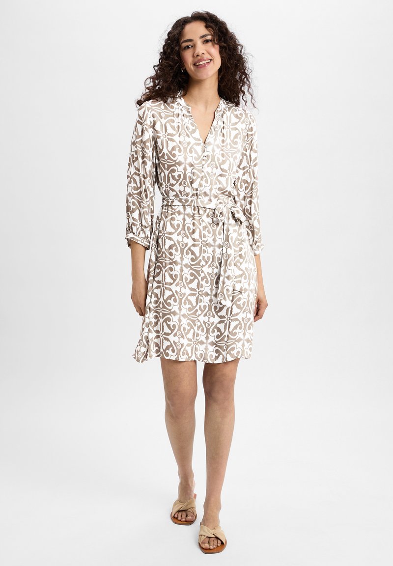 Short-sleeved beige dress with white intricate pattern, a collared neckline, belted waist, and a knee-length hem. Paired with tan sandals.