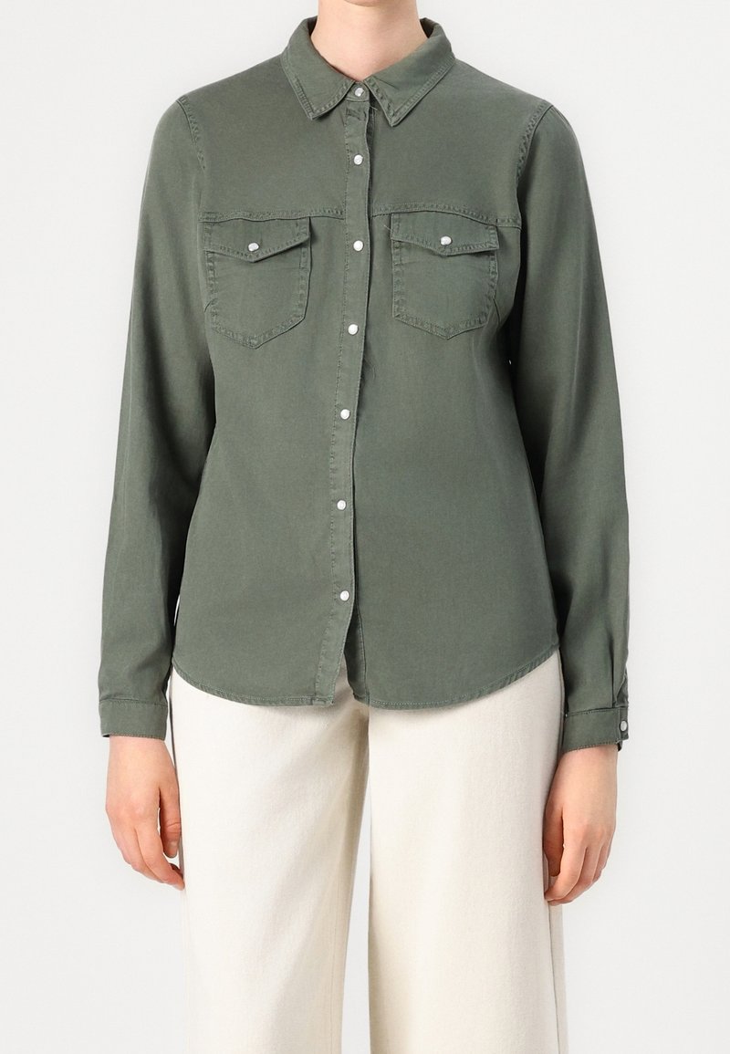 Personne portant une chemise kaki à manches longues boutonnée avec des poches à rabat et un pantalon large blanc cassé.