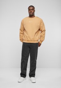Tan sweatshirt med rund hals og ribbede manchetter, med et logo på brystet, parret med mørkegrå bukser og hvide sneakers.