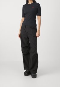 Czarna dopasowana koszulka z małym czerwonym logo, zestawiona z luźnymi czarnymi spodniami cargo i czarnymi klapkami. Stylizacja charakteryzuje się prostym, casualowym designem.