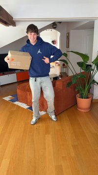 Pappkarton mit dem Text „Liebe mich. Trag mich.“, gehalten von einer Person in blauem Kapuzenpullover, hellen Jeans und silbernen Schuhen, mit einer Pflanze und einem orangefarbenen Sofa im Hintergrund.