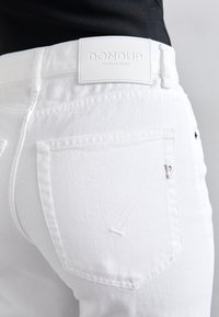 Balti džinsai su lygiu paviršiumi. Yra firminė balta odinė etiketė juosmens srityje, subtilus siuvinėjimas ir mažas metalinis logotipas ant galinio kišenės.