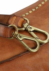 Angolo di una borsa in pelle marrone chiaro con due ganci girevoli in ottone attaccati a una tracolla marrone, decorata con rivetti in ottone lungo il bordo.