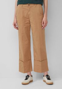 Pantalons larges couleur khaki en coton, avec une fermeture à bouton, des poches avant et un ourlet replié. Associés à des baskets crème.