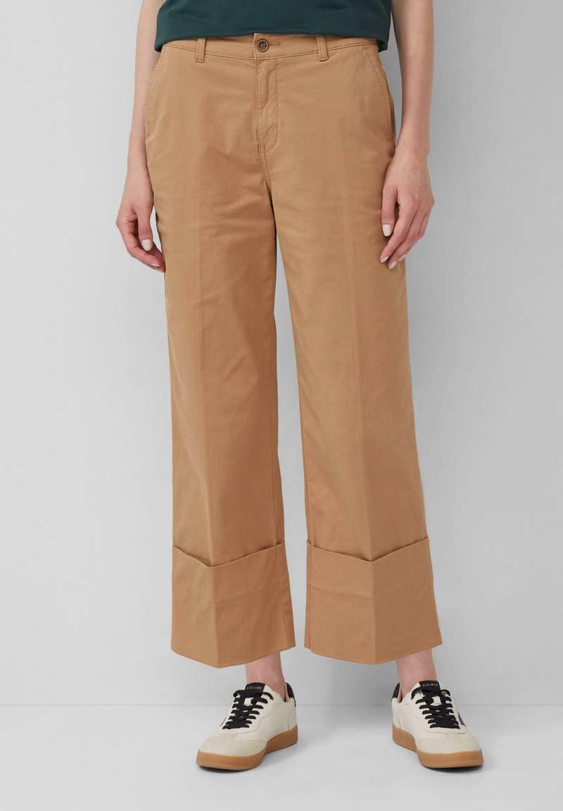 Pantalons larges couleur khaki en coton, avec une fermeture à bouton, des poches avant et un ourlet replié. Associés à des baskets crème.