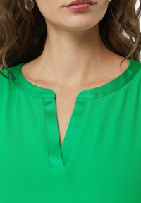 Green top with a V-neckline, made from smooth fabric. Features a clean, flat collar and a simple design with no visible patterns or embellishments.