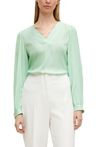 Blusa verde menta a maniche lunghe con scollatura a V e texture setosa. Presenta maniche arricciate ed è abbinata a pantaloni bianchi a vita alta.