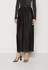 Černá plisovaná maxi sukně s jemnou texturou a elastickým pasem s logem. Nošená s černými sandály na tenké pásky.