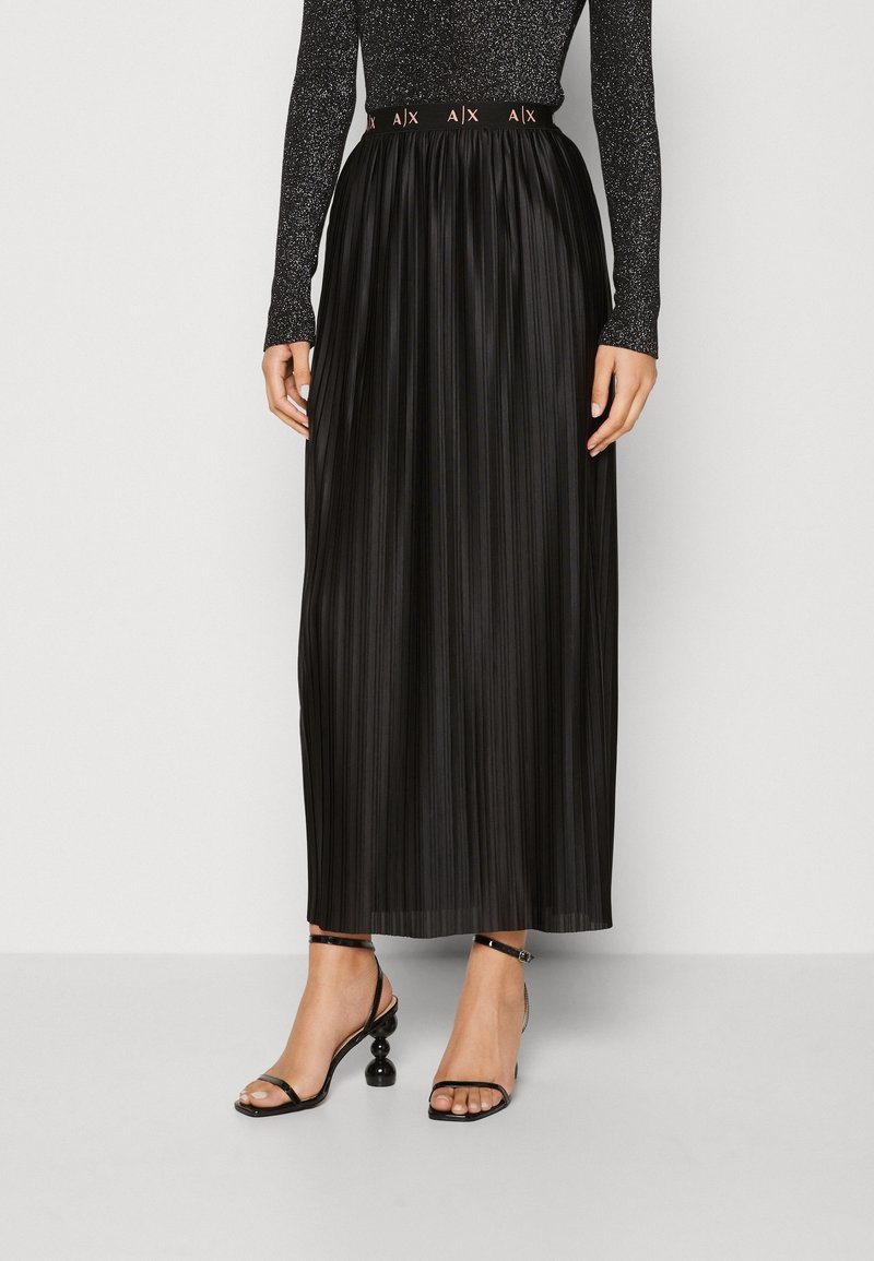 Černá plisovaná maxi sukně s jemnou texturou a elastickým pasem s logem. Nošená s černými sandály na tenké pásky.