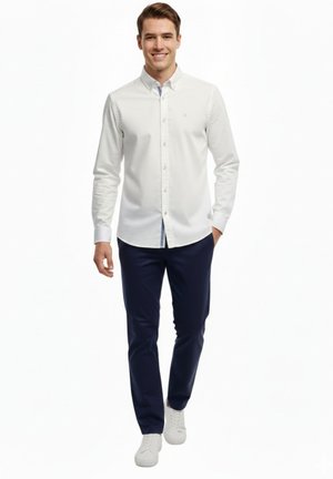 Hombre sonriente caminando, vestido con una camisa blanca abotonada, pantalones azul marino y zapatillas blancas, sobre un fondo blanco liso.