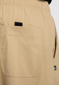 Beżowe szorty wykonane z lekkiego materiału z elastycznym paskiem w talii, wyposażone w boczną kieszeń, przeszycia dekoracyjne oraz małą metkę z logo.