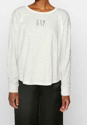 Person iført en hvid langærmet skjorte med "GAP" logo på brystet og sorte bukser, stående mod en ensfarvet lys baggrund.