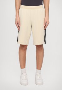 Pantalones cortos de algodón beige con una franja lateral negra, cintura elástica y dobladillo cosido, combinados con zapatillas blancas y calcetines claros.