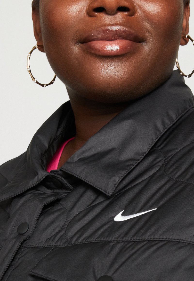 Veste rembourrée noire avec un col haut, présentant un logo Nike blanc et un intérieur contrastant rose. Des boucles d'oreilles en or circulaires sont visibles.