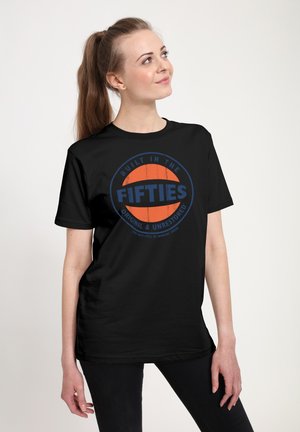 Jonge vrouw met paardenstaart en zwart T-shirt met blauw-oranje logo 'Built in the Fifties', staand tegen een effen lichte achtergrond.