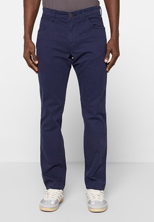 Mężczyzna ubrany w granatowe spodnie o prostych nogawkach i białe trampki, stojący na jednolitym jasnym tle.