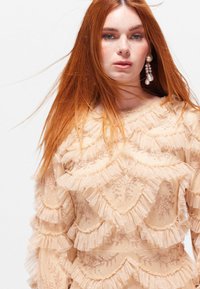 Pitkähihainen, läpikuultava beige mekko, jossa on kerrokselliset röyhelöt, kukkakirjonta ja teksturoitu kangas, sekä pyöreä pääntie.
