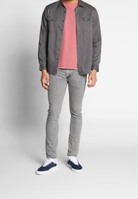 Grå skjorta med knappar över en rosa t-shirt, i kombination med ljusgrå jeans och marinblå sneakers med vita sulor, med en strukturerad design.