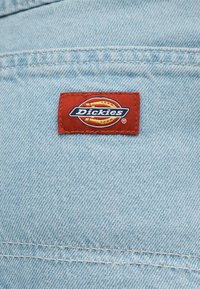 Κόκκινη ορθογώνια ετικέτα της μάρκας Dickies ραμμένη σε ανοιχτό γαλάζιο τζιν ύφασμα με εμφανείς οριζόντιες και κάθετες ραφές.