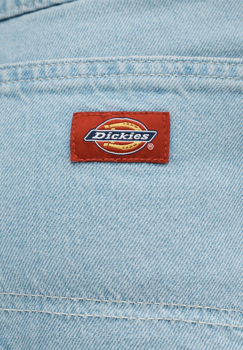 Rotes rechteckiges Dickies-Markenlabel auf hellblauem Denimstoff mit sichtbaren horizontalen und vertikalen Nähten aufgenäht.