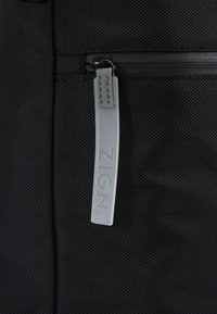 Gros plan d’un tissu noir texturé avec une fermeture éclair comportant une tirette translucide étiquetée "ZIGN" en lettres en relief.