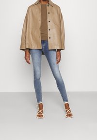 Chaqueta de cuero marrón claro, top marrón, jeans ajustados azul claro y sandalias blancas con tiras. Las características incluyen un bolsillo y cierre de botón en la chaqueta.