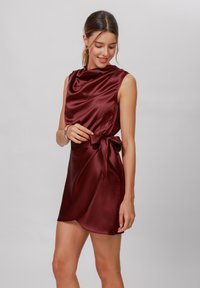 Burgundy satin dress with a cowl neckline, sleeveless design, and a tie waist, featuring a draped front and a knee-length hemline.