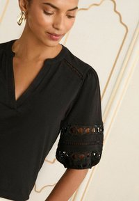 Juoda bluzelė su V formos iškirpte, pūstomis rankovėmis, puoštomis sudėtingomis išpjovų ornamentais, ir nėrinių detalėmis ant pečių. Minkšta audinio tekstūra.