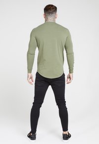 Langärmliges olivgrünes Shirt mit abgerundetem Saum, getragen zu schmal geschnittenen schwarzen Jeans und schwarzen Sneakers mit weißen Sohlen. Glatte Stofftextur.