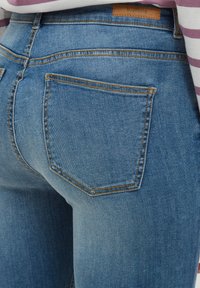 Modré džíny z denimu s detailním pohledem na zadní stranu, zobrazující dva kapsy s oranžovým prošíváním a béžovou nášivkou na pase.