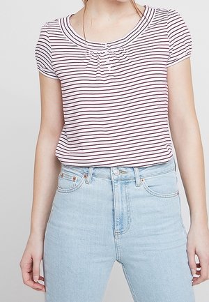 Frau trägt ein weißes T-Shirt mit kurzen Ärmeln und dünnen schwarzen horizontalen Streifen und eine hellblaue Jeans mit hoher Taille vor einem grauen Hintergrund.