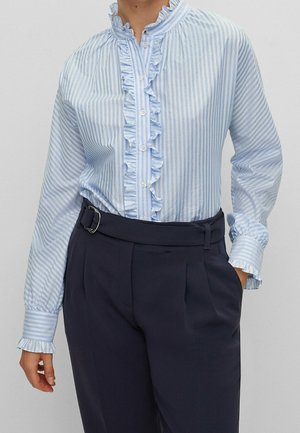 Femme portant un chemisier rayé vertical bleu clair et blanc avec col et poignets à volants, rentré dans un pantalon taille haute bleu marine avec ceinture.